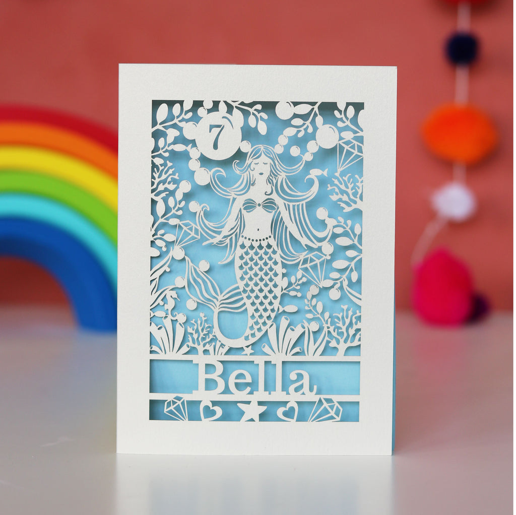 A cream and light blue laser cut birthday card with a mermaid and a name and age. 
