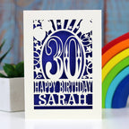 A cream personalised paper cut birthday card cut away to reveal a violet background. Cut design is an age surrounded by stars and balloons, with the words "Happy birthday" and a name underneath