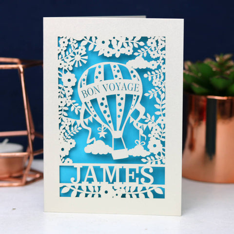 Beautiful papercut cream card showing a hot air balloon  with "Bon Voyage".  Bright blue insert paper , sun and flowers and the name James across the bottom.