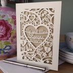 A Personalised Papercut wedding anniversary Card featuring the words "Mr and Mrs Stevens" and the date inside a heart, surrounded by flowers and leaves. There are two hearts underneath with an initial in each heart. 
