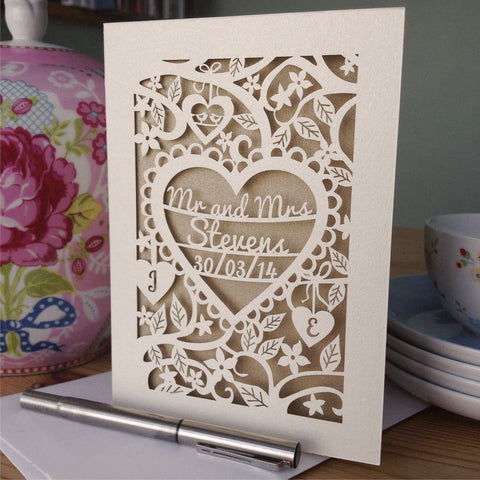 A Personalised Papercut wedding Card featuring the words "Mr and Mrs Stevens" and the date inside a heart, surrounded by flowers and leaves. There are two hearts underneath with an initial in each heart.  - A5 / Gold Leaf