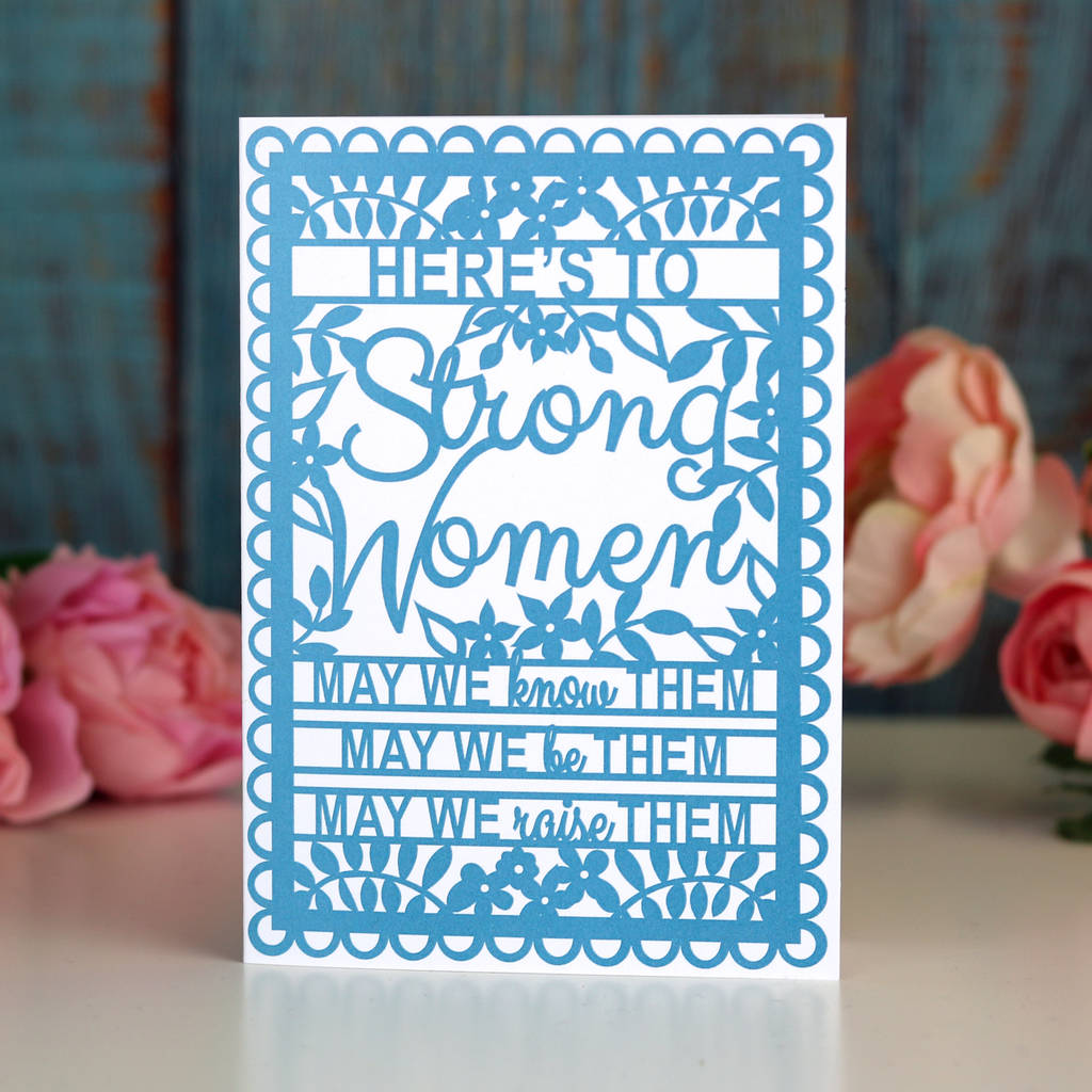 A blue printed greeting card with the text 'HERE'S TO STRONG WOMEN' and a floral pattern around the border, placed on a wooden surface with flowers in the background.