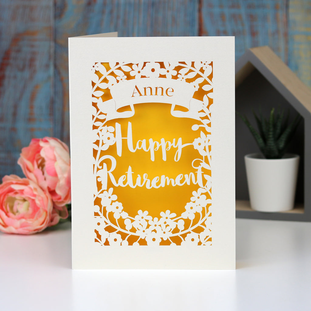 Laser cut personalised retirement card. Cut from cream card with a sunshine yellow insert paper. Shows the words "Happy Retirement" surrounded by flowers and a banner for the personalisation.
