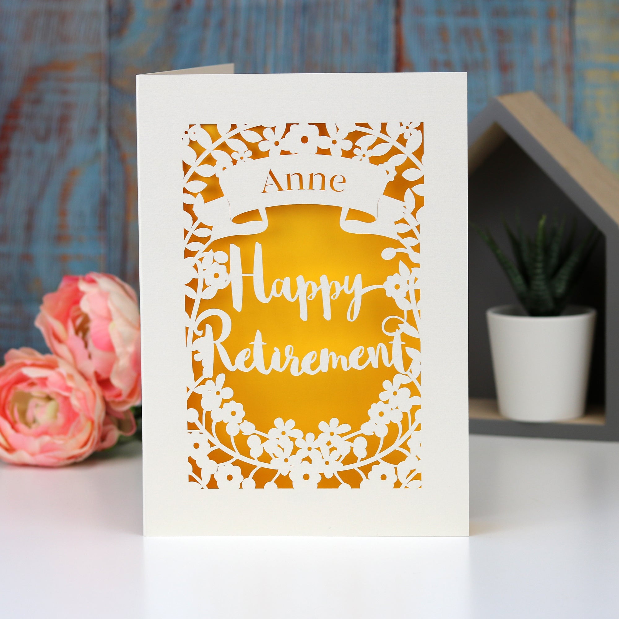 Laser cut personalised retirement card. Cut from cream card with a sunshine yellow insert paper. Shows the words "Happy Retirement" surrounded by flowers and a banner for the personalisation.